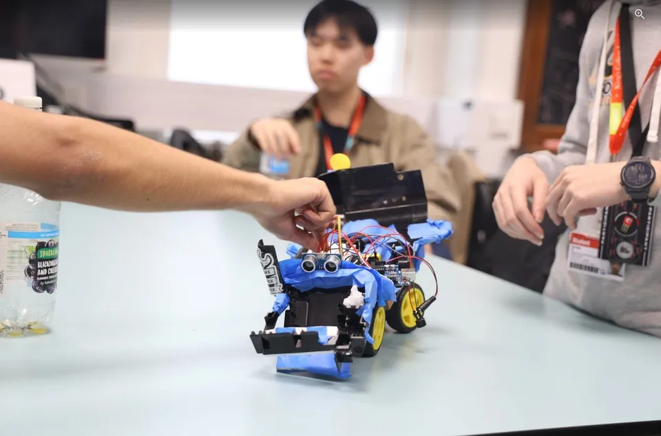 A hand adjusts a small wheeled robot wrapped in blue tape, built from salvaged parts