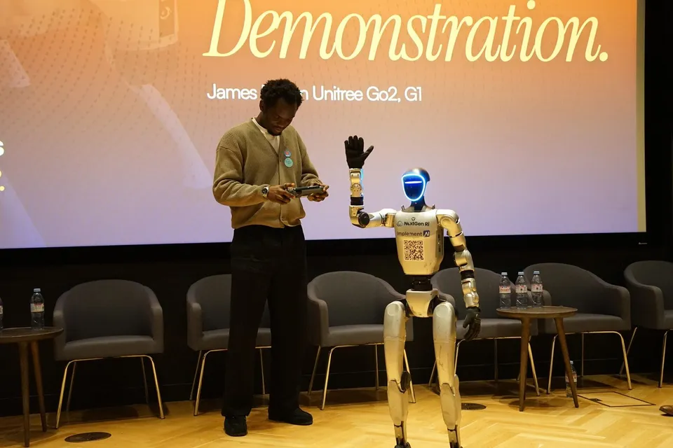 A Unitree G1 humanoid robot raises its hand on stage beside a presenter at Tomorrow’s Intelligence